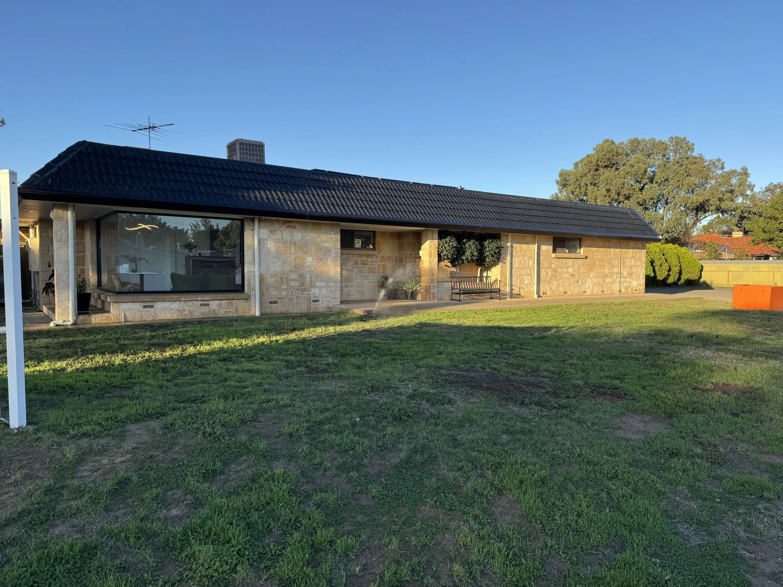 A single-story house with a black tiled roof, stone exterior walls, a large front window, and a small window. There is a front lawn with patches of dirt, a bench, a small tree, a sprinkler, and some bushes. The sky is clear and blue with a few trees in the background.