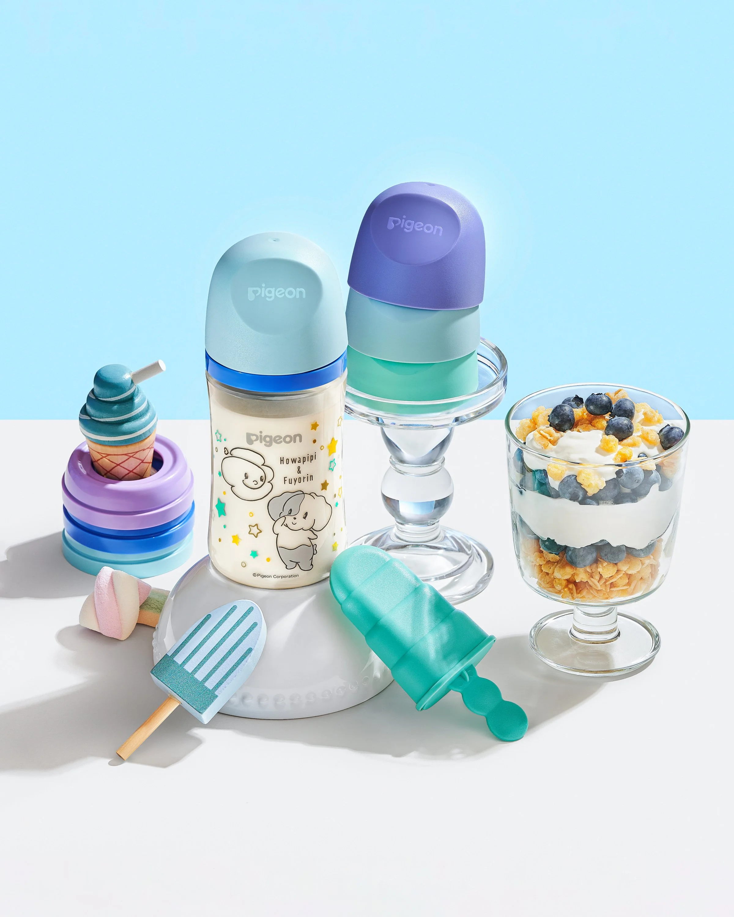 Stacked colorful baby bottles, a glass of yogurt with blueberries and cereal, ice cream-shaped toy, and toy spatulas on a white table with a blue background.