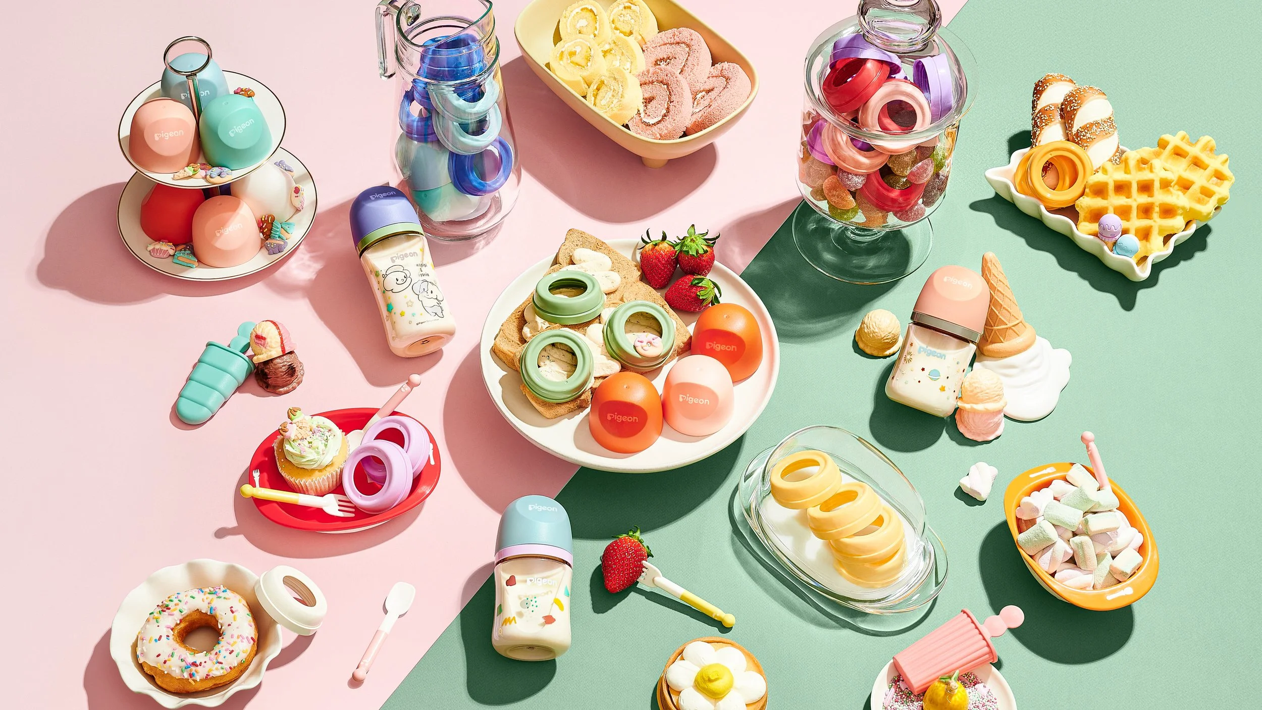 Colorful assortment of toy pretend foods and beverages arranged on pastel pink and green surfaces. Includes desserts like donuts, waffles, marshmallows, and candies, as well as toy bottles, jars, and fruit slices, all designed for children's play.
