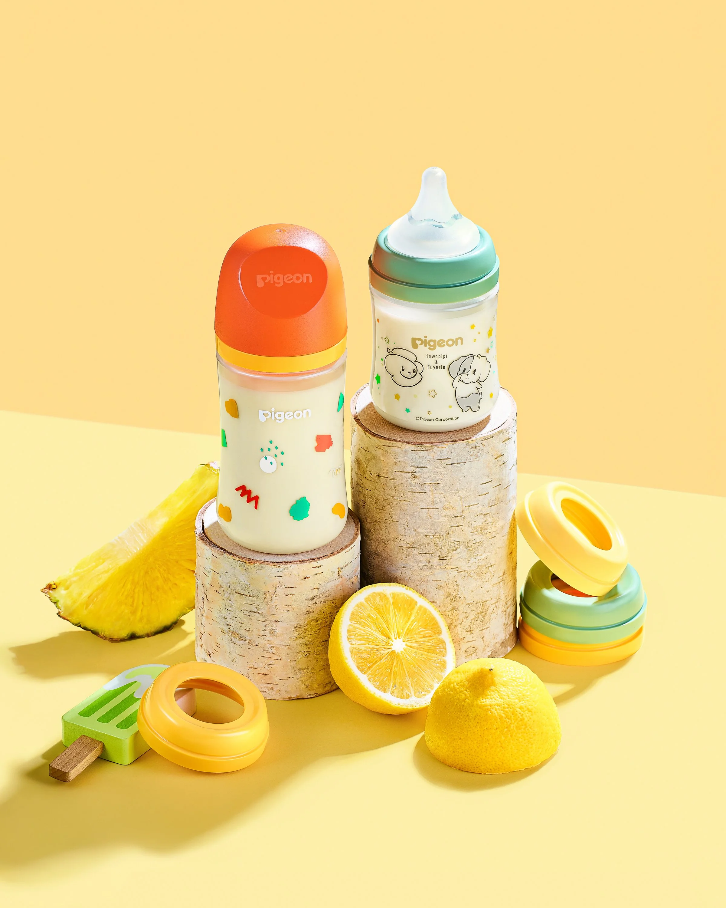 Two baby bottles with colorful caps and illustrated designs placed on wooden stumps, with pineapple, lemon, and lemon half, and stacking rings toy on a yellow background.