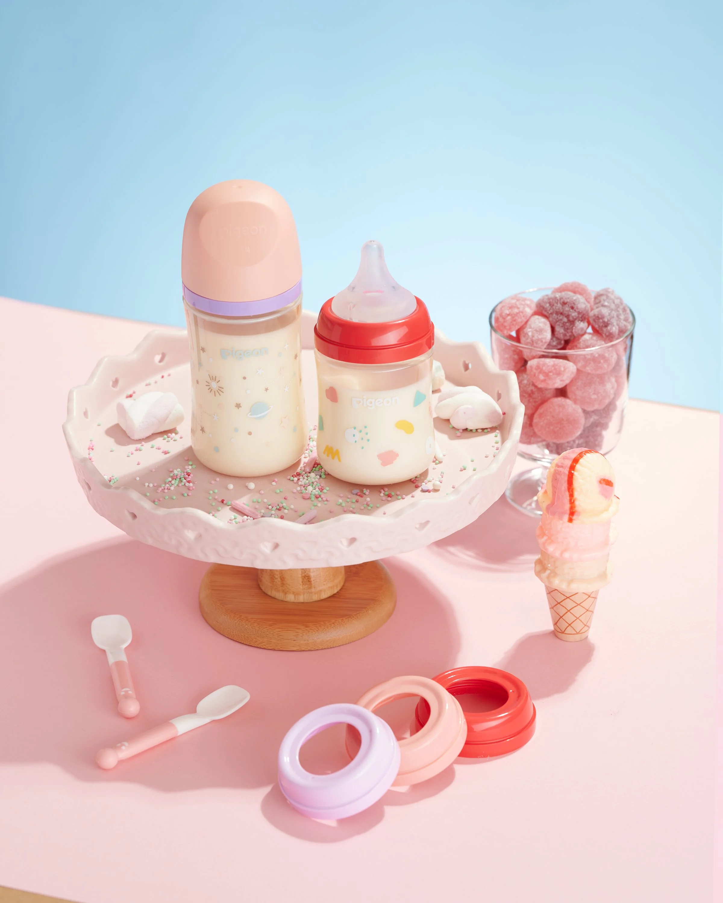 Two baby bottles with pastel-colored caps and designs, a glass of pink candies, a small ice cream cone, and a set of plastic teething rings and utensils on a pink and blue background.