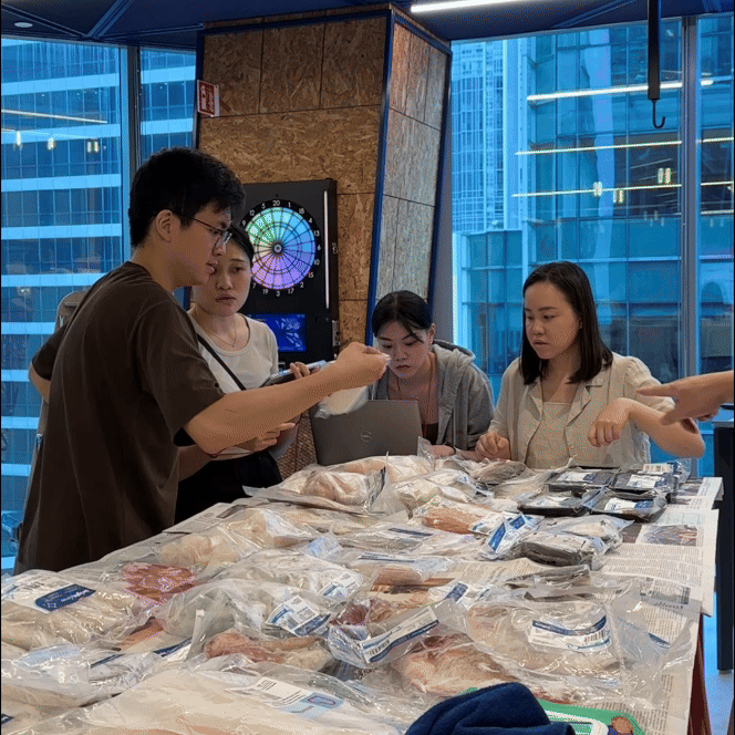 Four women and one man examining packaged meat on a table at a market or store, with a dartboard and large windows in the background.