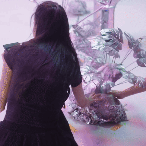 A person with long dark hair, wearing a black dress, reaching towards plants wrapped in metallic-colored foil.