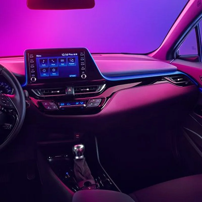 The interior of a modern car dashboard illuminated in pink and purple lighting, featuring a digital touchscreen display, climate controls, and gear shift lever.