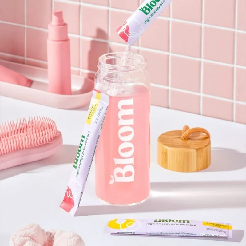 A pink bottle labeled 'Bloom' with a packet of high energy pre-workout supplement being poured into it, on a white bathroom counter with a pink tiled wall in the background.