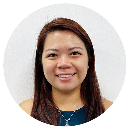 A young woman with straight, shoulder-length brown hair, smiling, wearing a dark top and a necklace with a star pendant, standing against a plain white wall.