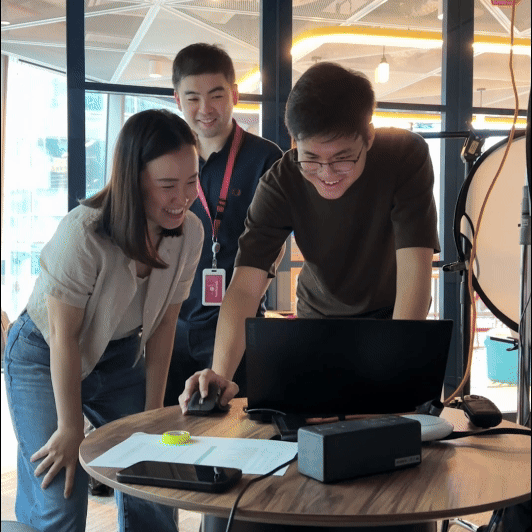 Three people smiling and looking at a laptop on a table in a modern indoor space.