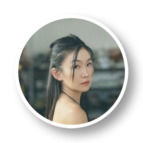 A young woman with long dark hair tied back, looking over her shoulder, with an indoor background.