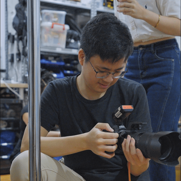 Young man wearing glasses, sitting and looking at a camera with a large telephoto lens, in a room with shelves of equipment.
