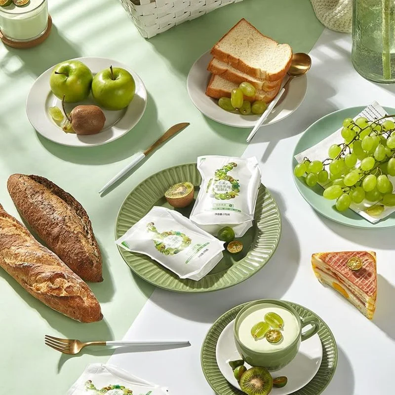 A breakfast table with green and white tableware, including apples, grapes, bread, sliced bread, a slice of cake, kiwi, a green drink, and packaged snacks.