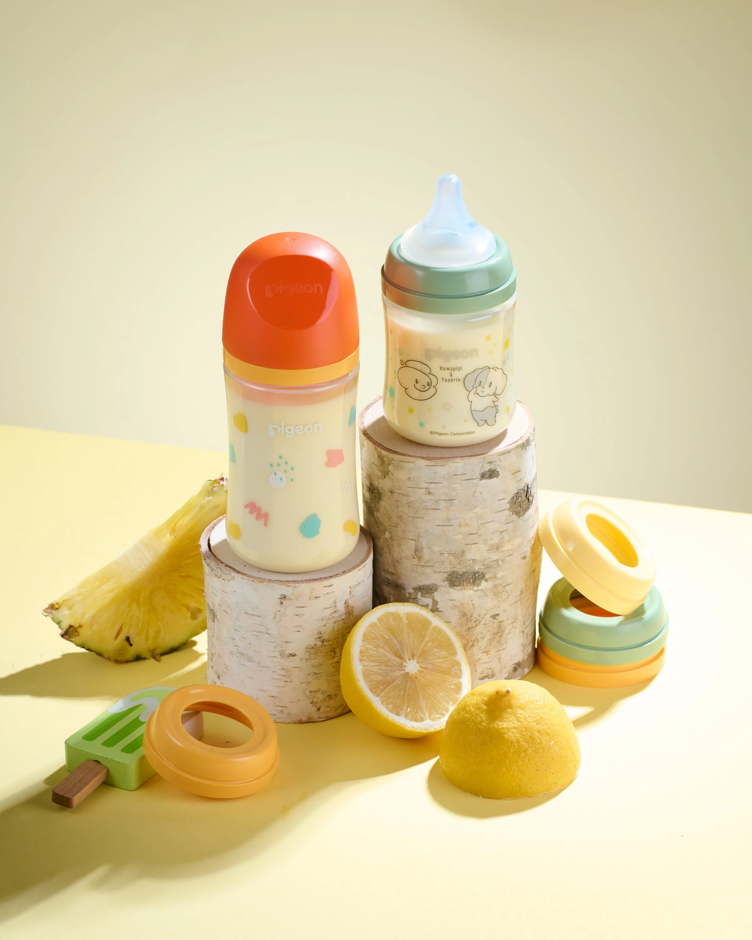 Two baby bottles with pastel-colored caps and illustrations, pineapples and lemons, and bamboo stands on a light yellow background.