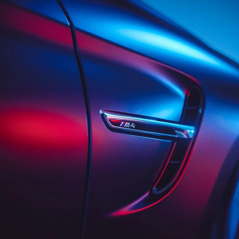 Close-up of a BMW M4 badge on the side of a car, illuminated by blue and red lighting.