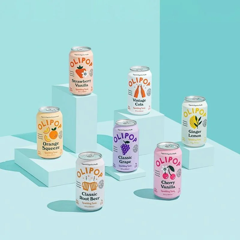 Seven cans of Olipop sparkling tonic arranged on light blue risers with a pastel blue background. The flavors include Strawberry Vanilla, Vintage Cola, Ginger Lemon, Orange Squeeze, Classic Grape, Cherry Vanilla, and Classic Root Beer.