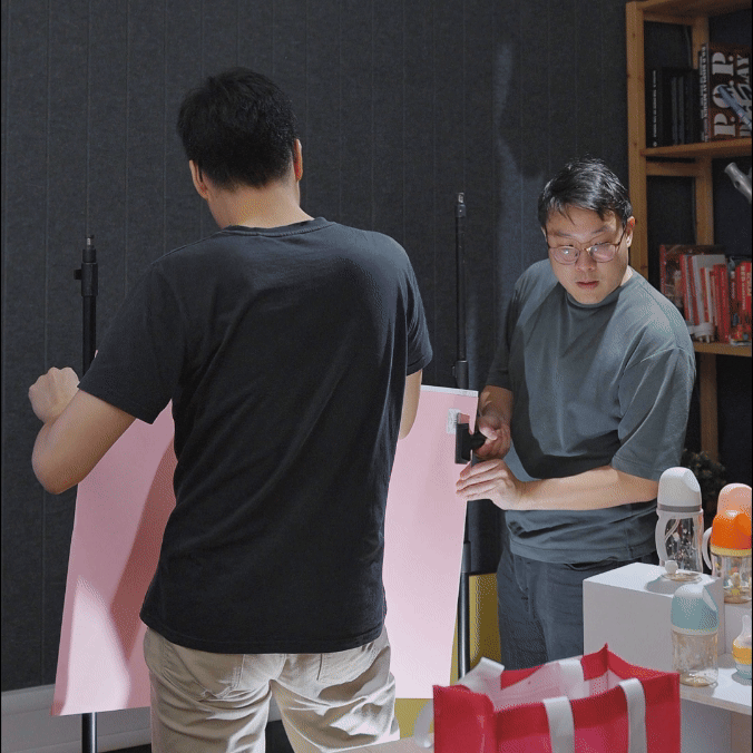 Two men setting up a pink backdrop in a room with bookshelves and various bottles on a table.