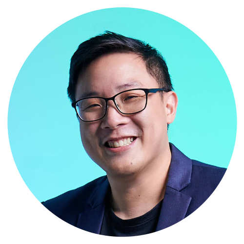 Portrait of a young man with glasses smiling, wearing a dark blazer against a light blue background.