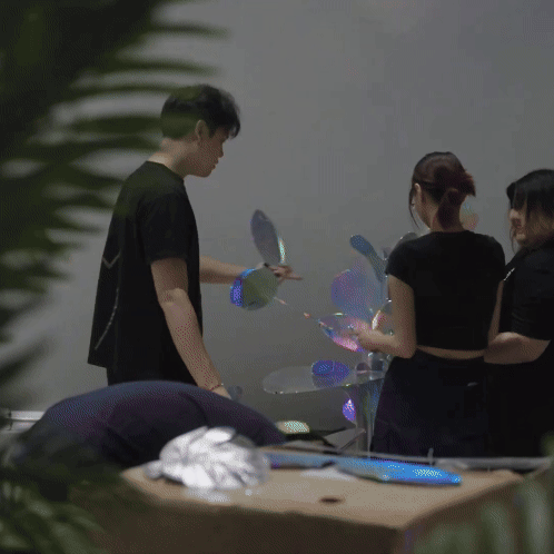 Three people standing indoors, with one person in front and two others behind, seemingly involved in a demonstration or activity involving reflective, holographic disks.