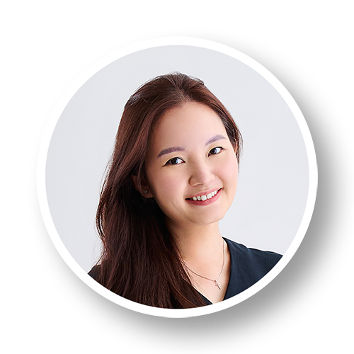 Smiling young woman with long brown hair, wearing a navy top and a necklace, against a white background.