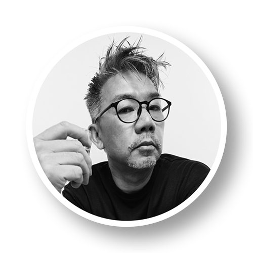Black and white portrait of a man with spiky hair and round glasses, making a gesture with his hand.