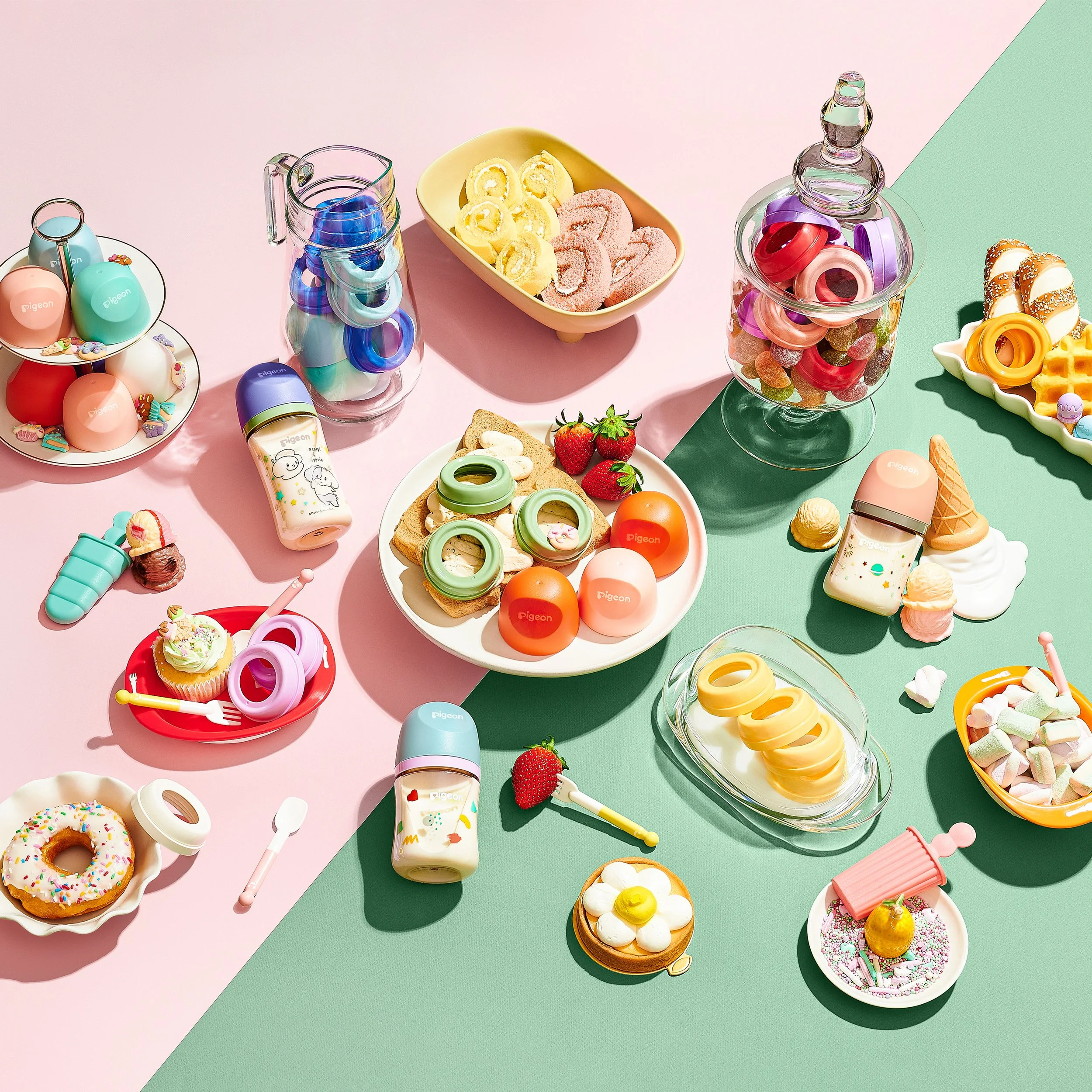 An assortment of colorful toy desserts, drinks, and plates arranged on a pastel pink and green split background, resembling a tea party setup.