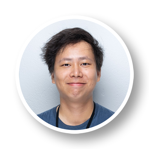 Close-up portrait of a smiling young man with dark hair wearing a blue shirt against a light gray background.