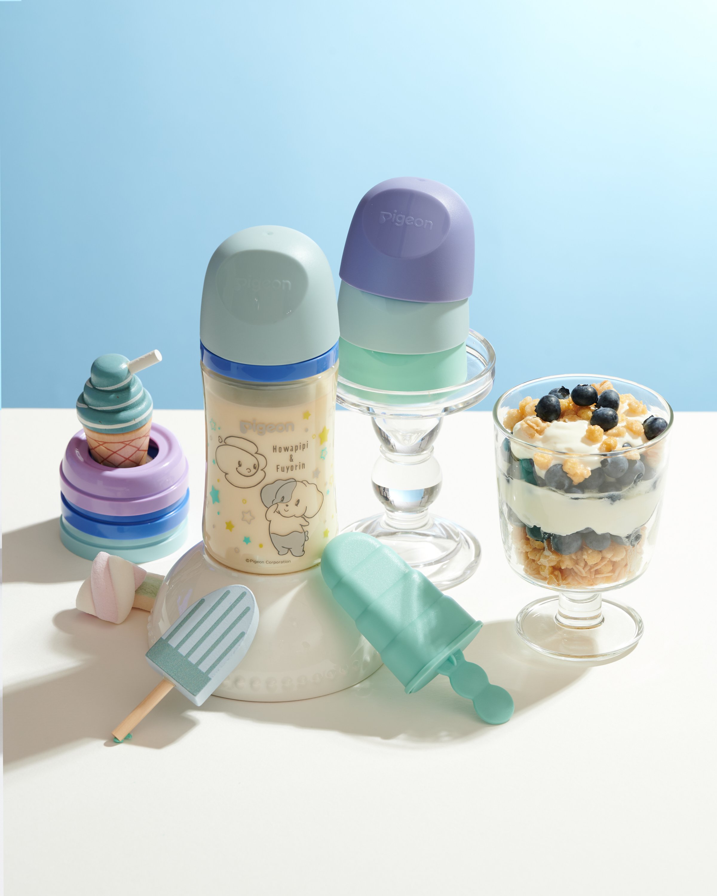 Stacked pastel-colored baby bottles, two ice cream-shaped toys, and a parfait glass with layers of blueberries, cereal, and yogurt on a white surface with a light blue background.