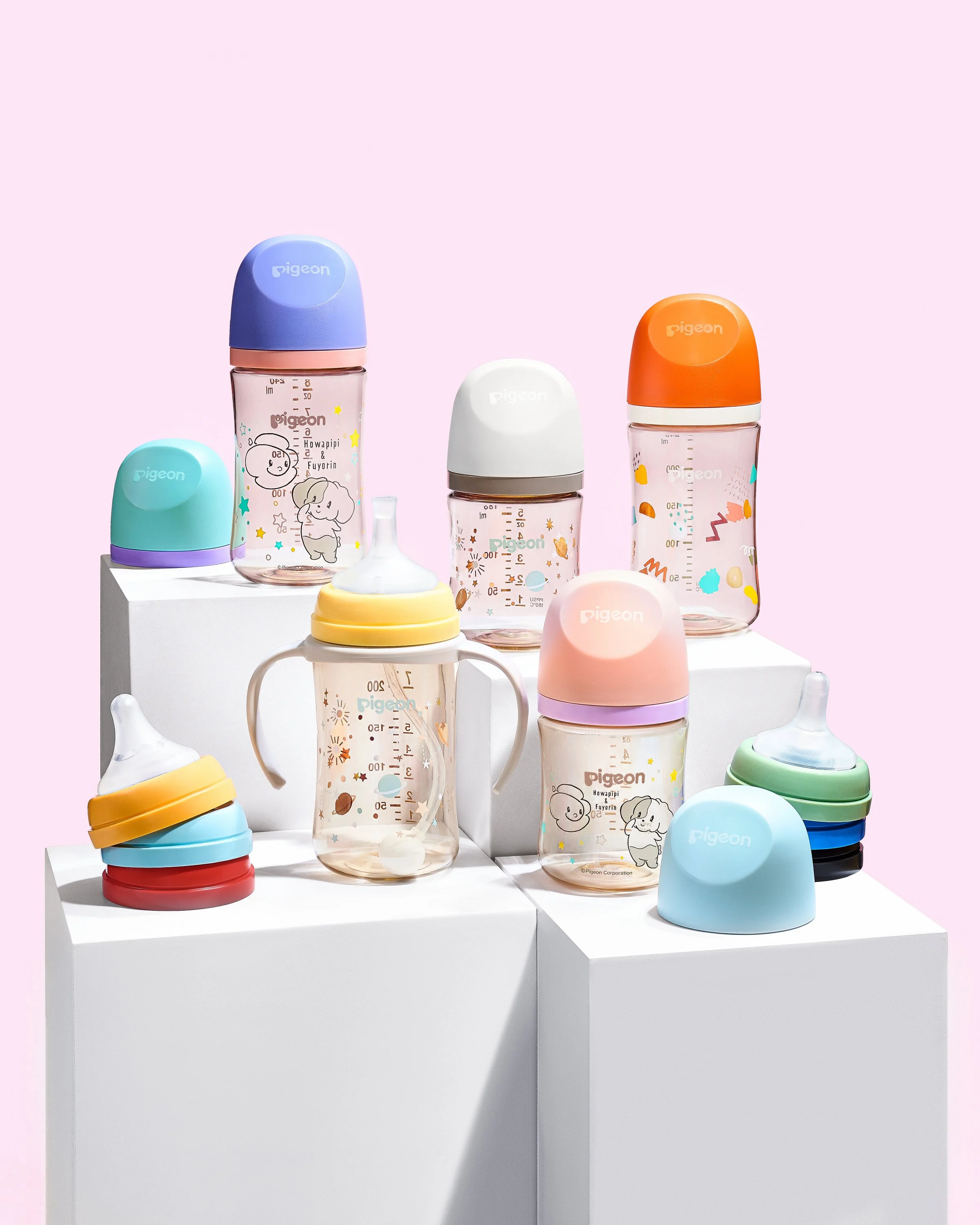 Collection of colorful baby bottles and pacifiers displayed on white pedestals against a pink background.