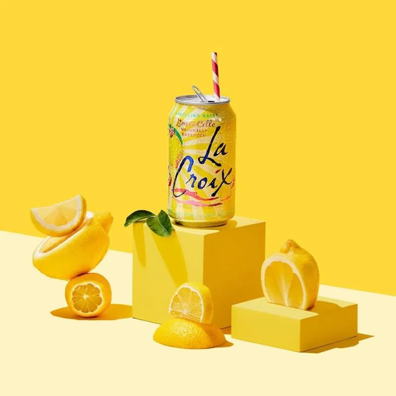 A can of La Croix lemon-flavored sparkling water with a pink and white striped straw, surrounded by lemon slices, lemon wedges, and a green leaf, all set against a yellow background with yellow platforms.