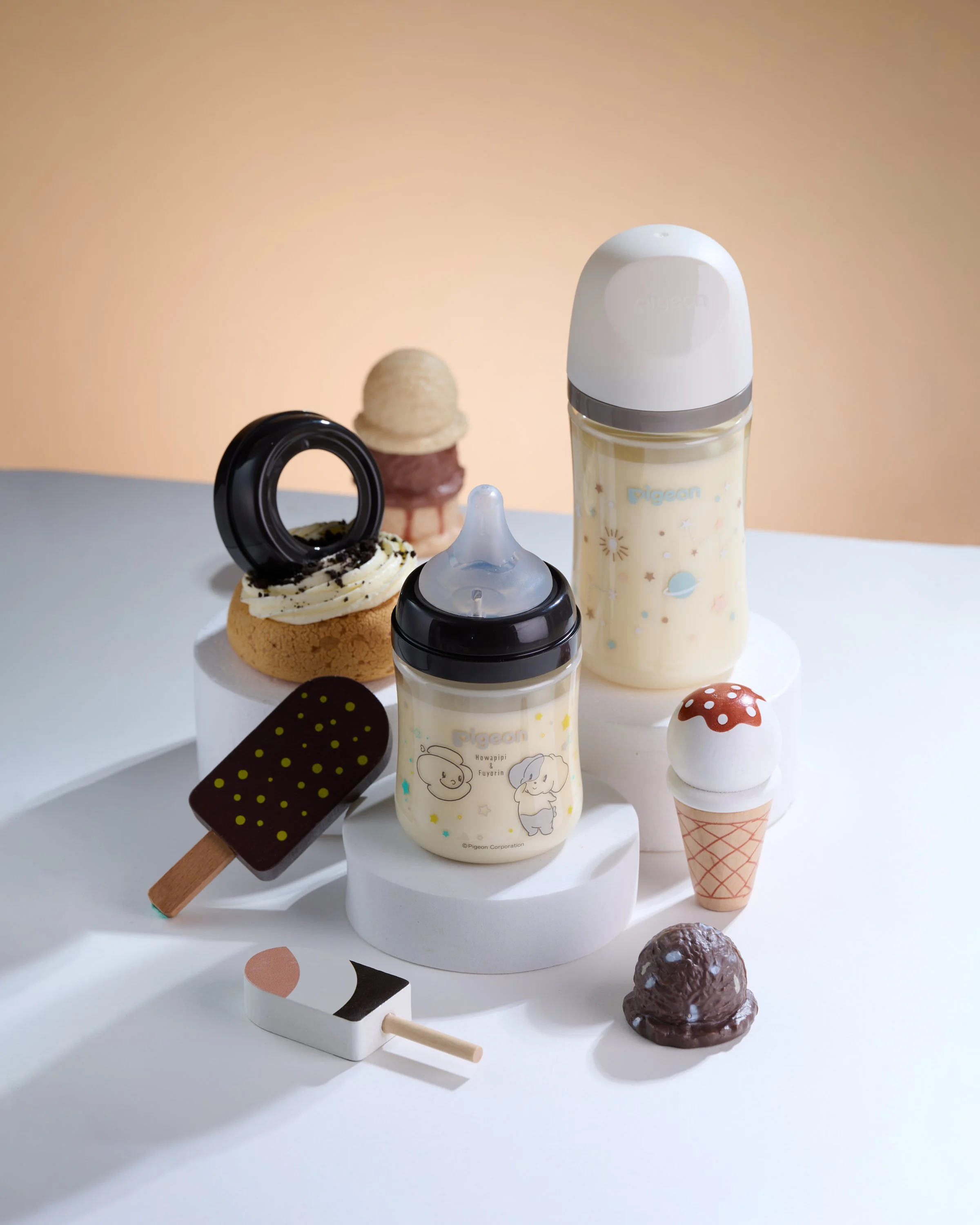 Collection of baby bottles, toy ice cream, and toy food replicas on a white surface with a beige background.