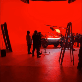 Behind-the-scenes photo of a car reveal event with a red backdrop, a black car with glowing headlights, and people observing, with studio equipment and ladders present.