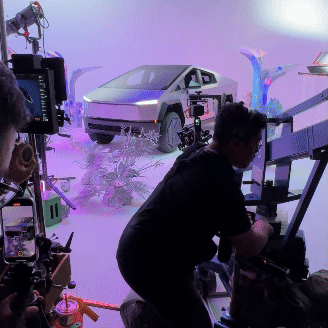 A scene from a behind-the-scenes film or photo shoot featuring a futuristic car model on a set with purple and blue lighting, with crew members operating equipment.
