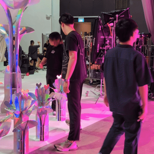 People setting up or working at a colorful, modern exhibition or display area with iridescent plants and equipment bathed in pink and purple lighting.