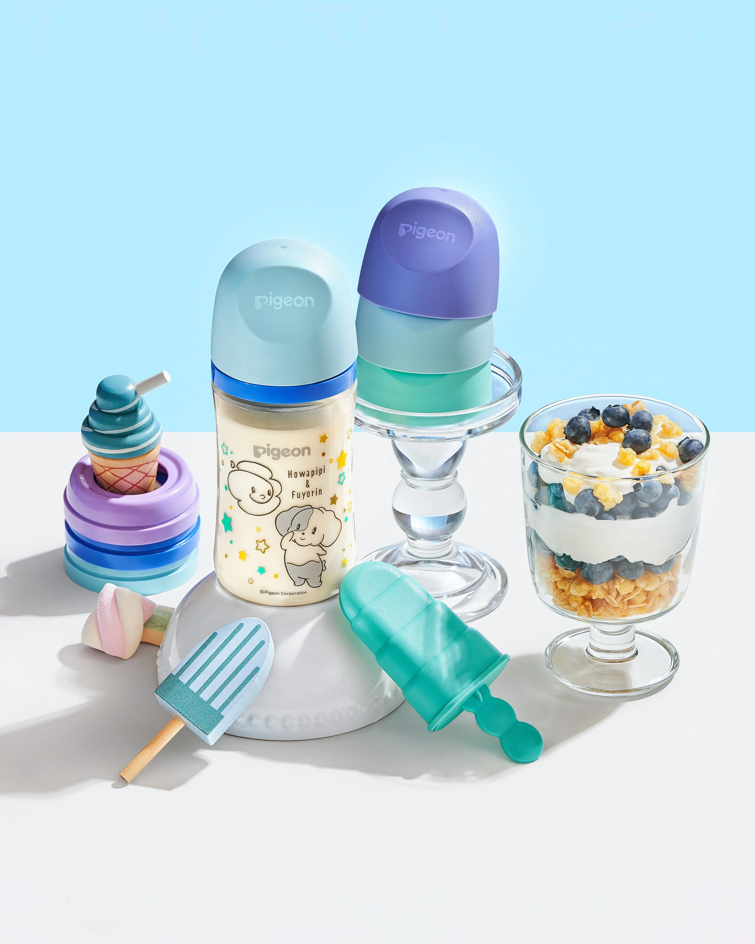 Colorful children's ice cream toy set with plastic ice cream cones, popsicles, and a dessert in a glass cup, arranged on a white table with a blue background.