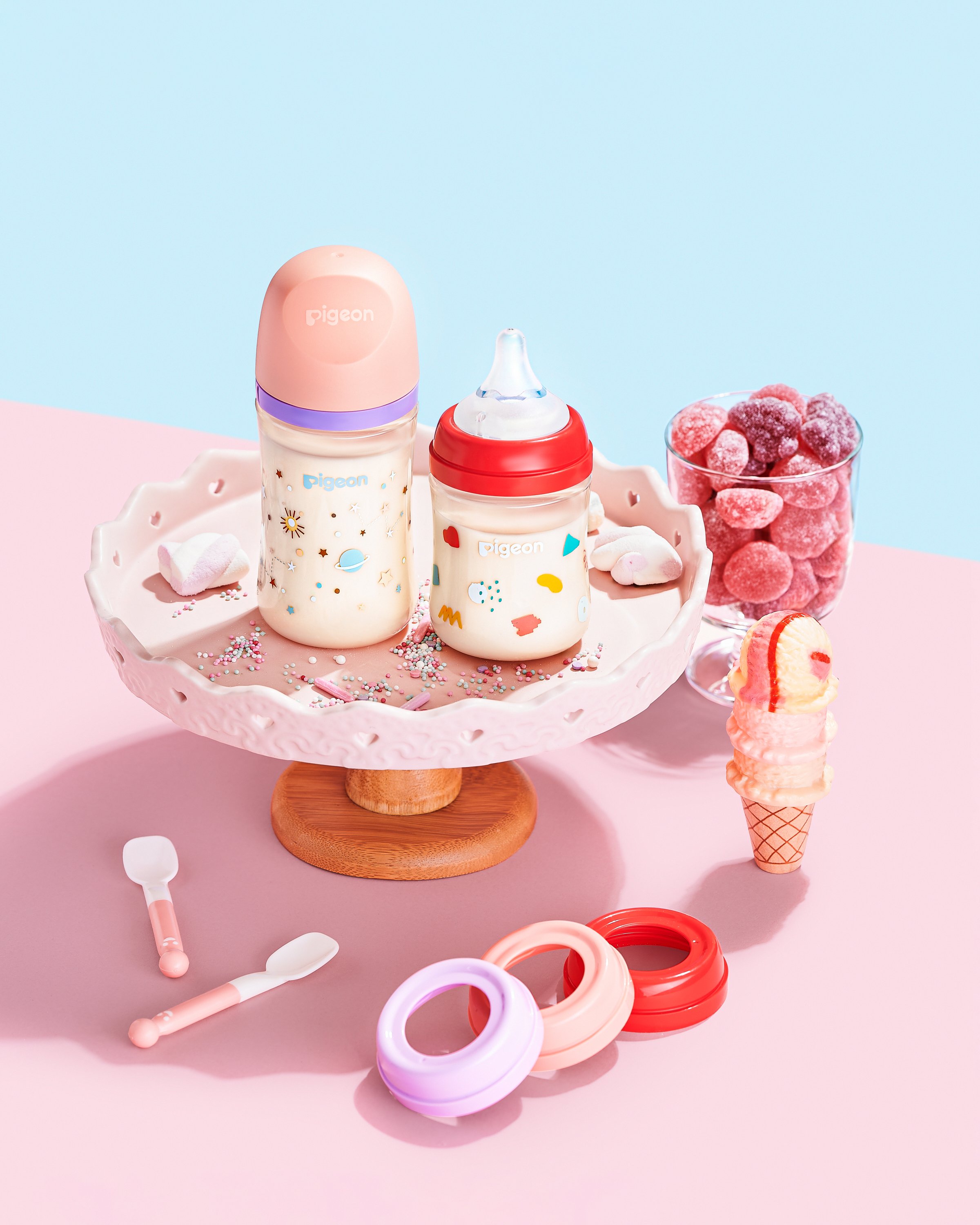 Two baby bottles with colorful caps and designs on a pink cake stand, surrounded by a cup of pink candies, an ice cream cone, and various baby accessories, set on a pink and blue background.