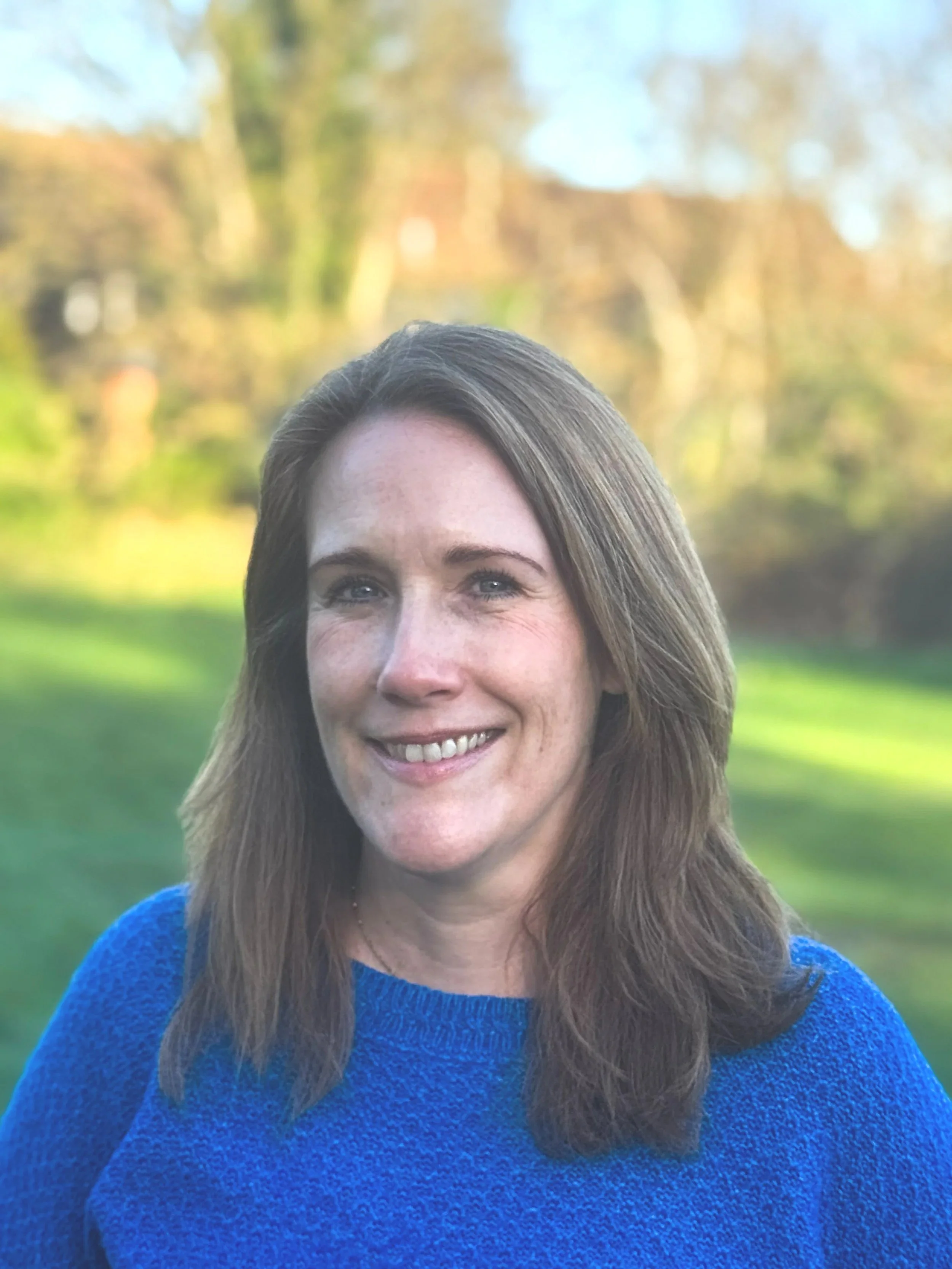 A woman with shoulder-length brown hair wearing a blue sweater, smiling outdoors with a blurred background of trees and grass.