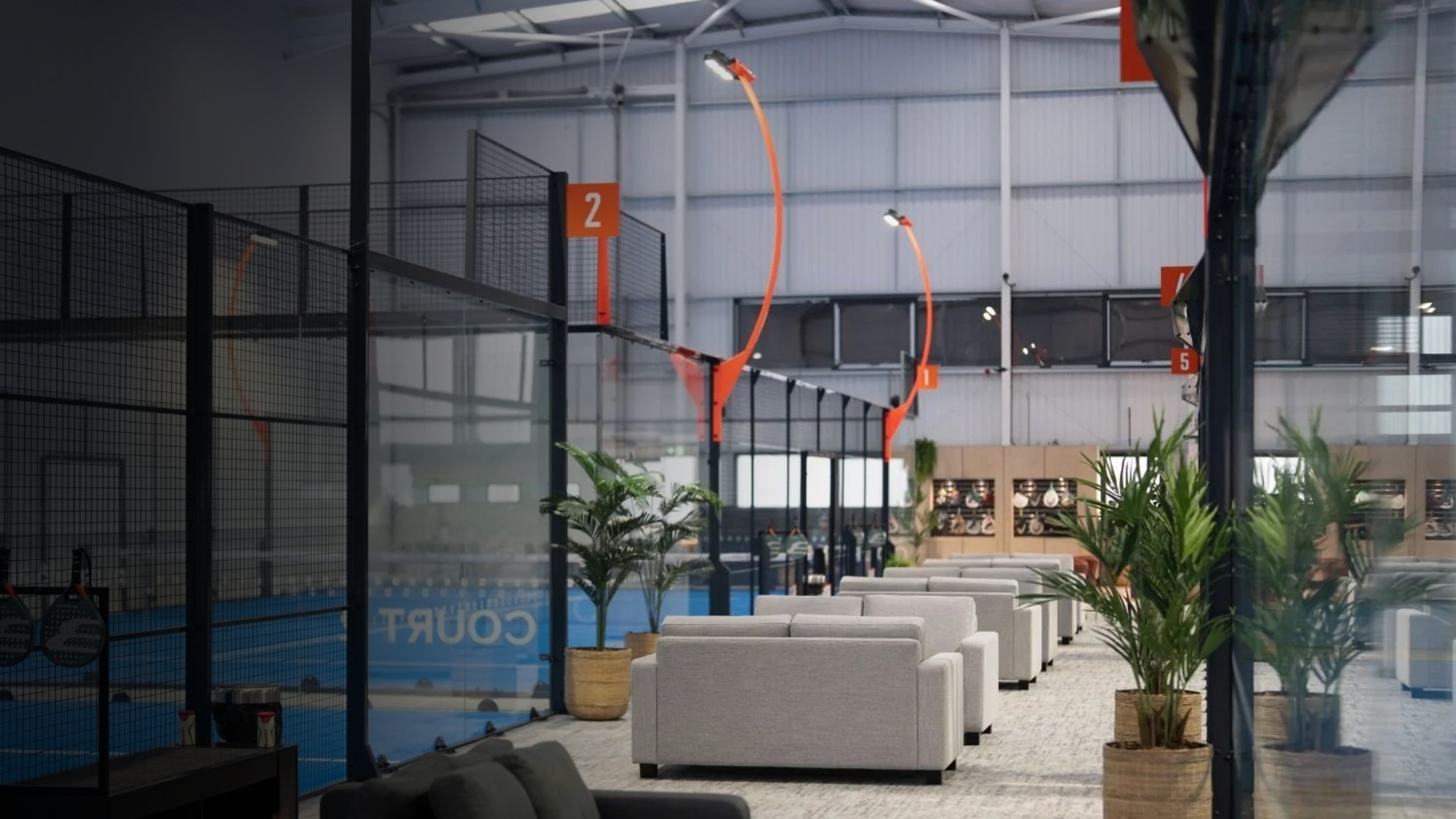 Indoor sports facility with padel courts, seating area with white sofas, and large potted plants.