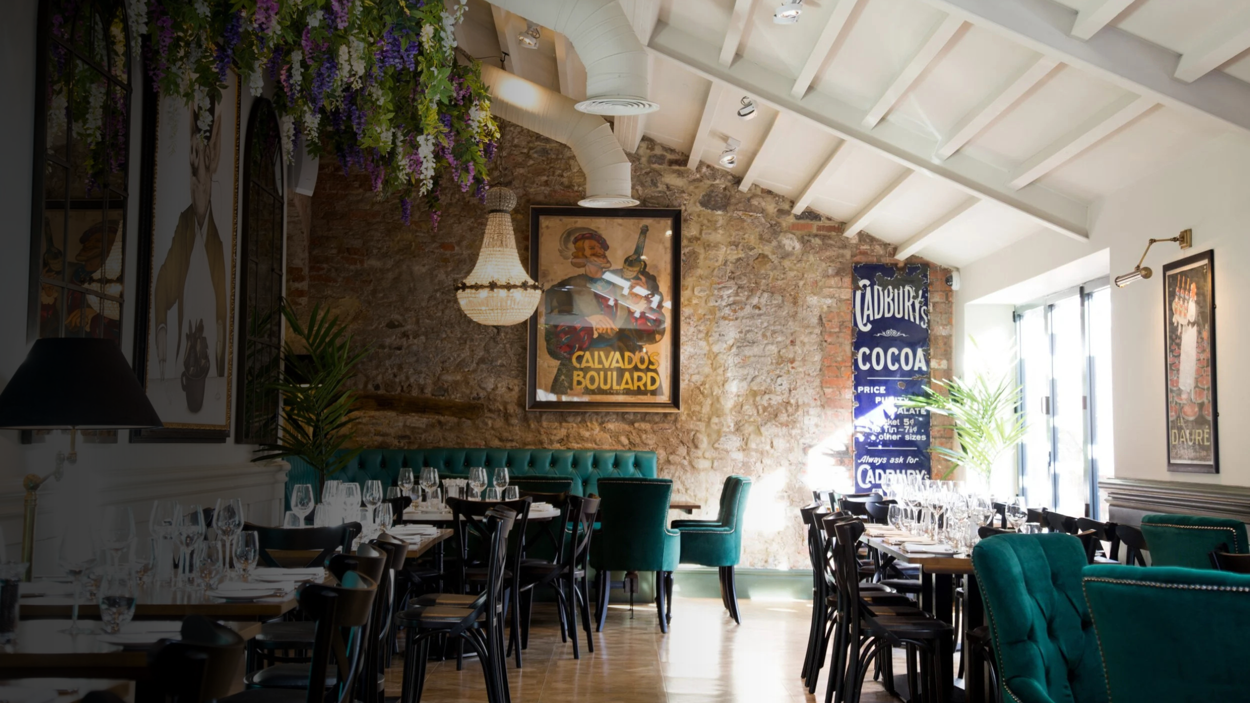 Interior of a restaurant with tables set for dining, featuring wine glasses and napkins. The space has an exposed brick wall with vintage posters, a chandelier, and a large window letting in natural light. There are green velvet chairs and plants adding to the decor.