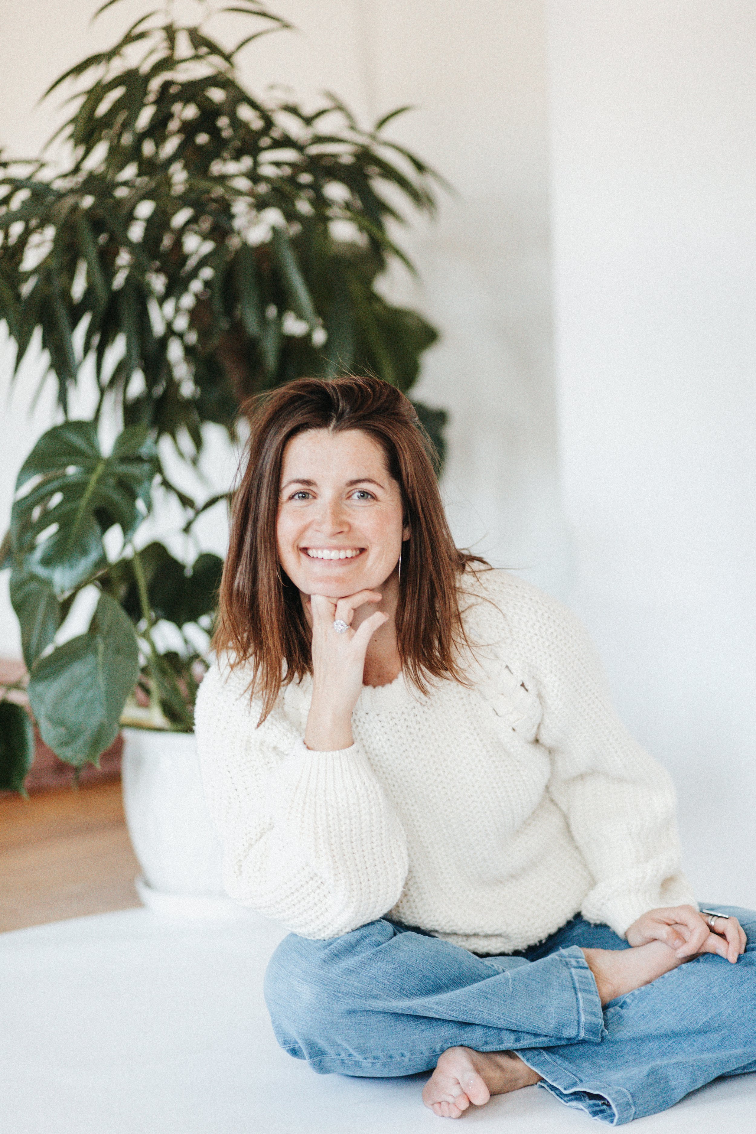 A woman with shoulder-length brown hair and freckles, smiling, sitting cross-legged on a white surface, wearing a cream sweater and blue jeans, with a large leafy plant in the background.