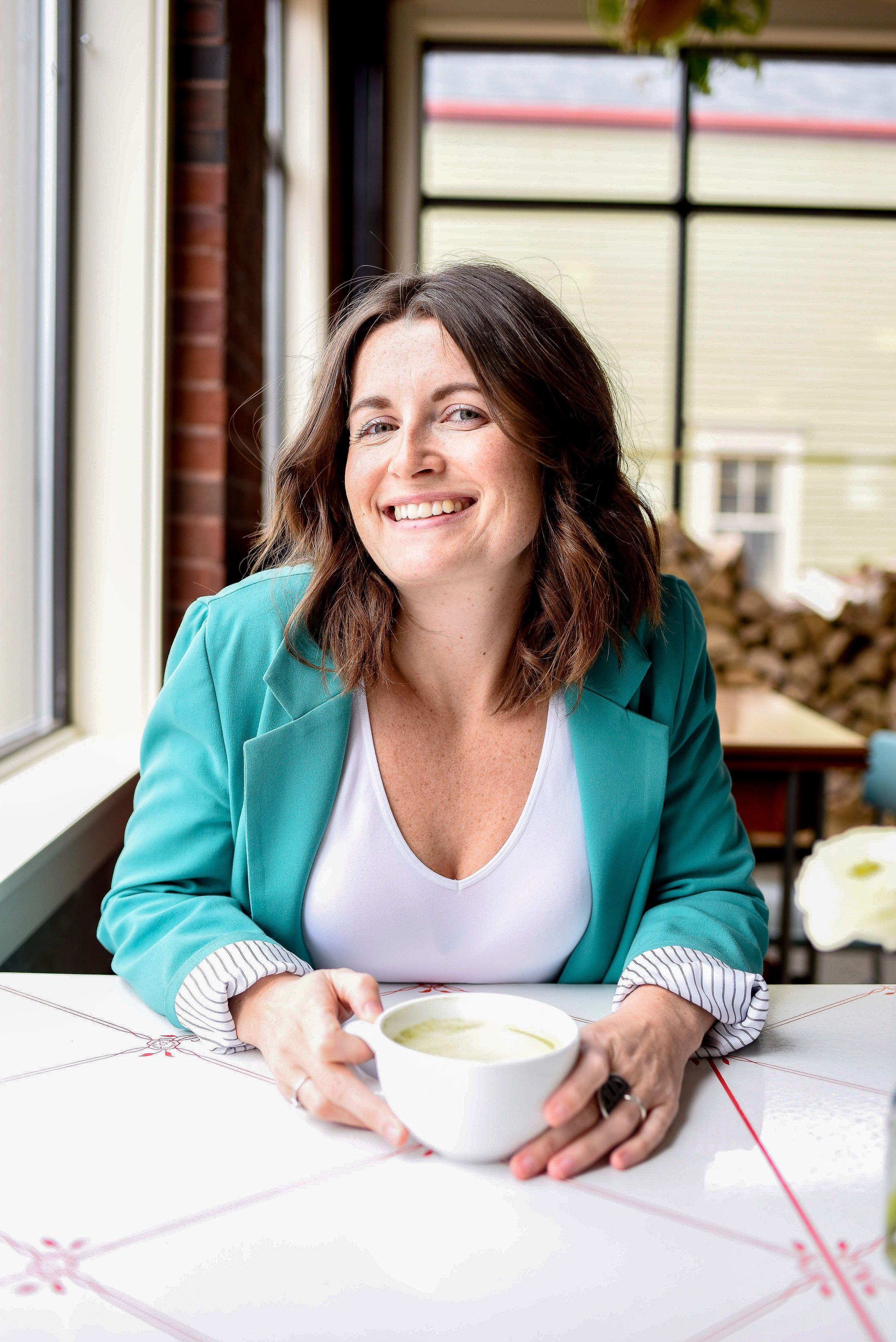 A woman with shoulder-length brown hair, wearing a teal blazer over a white top, smiling while holding a white mug with a green beverage inside, seated at a table near large windows with a brick wall and house outside in the background.