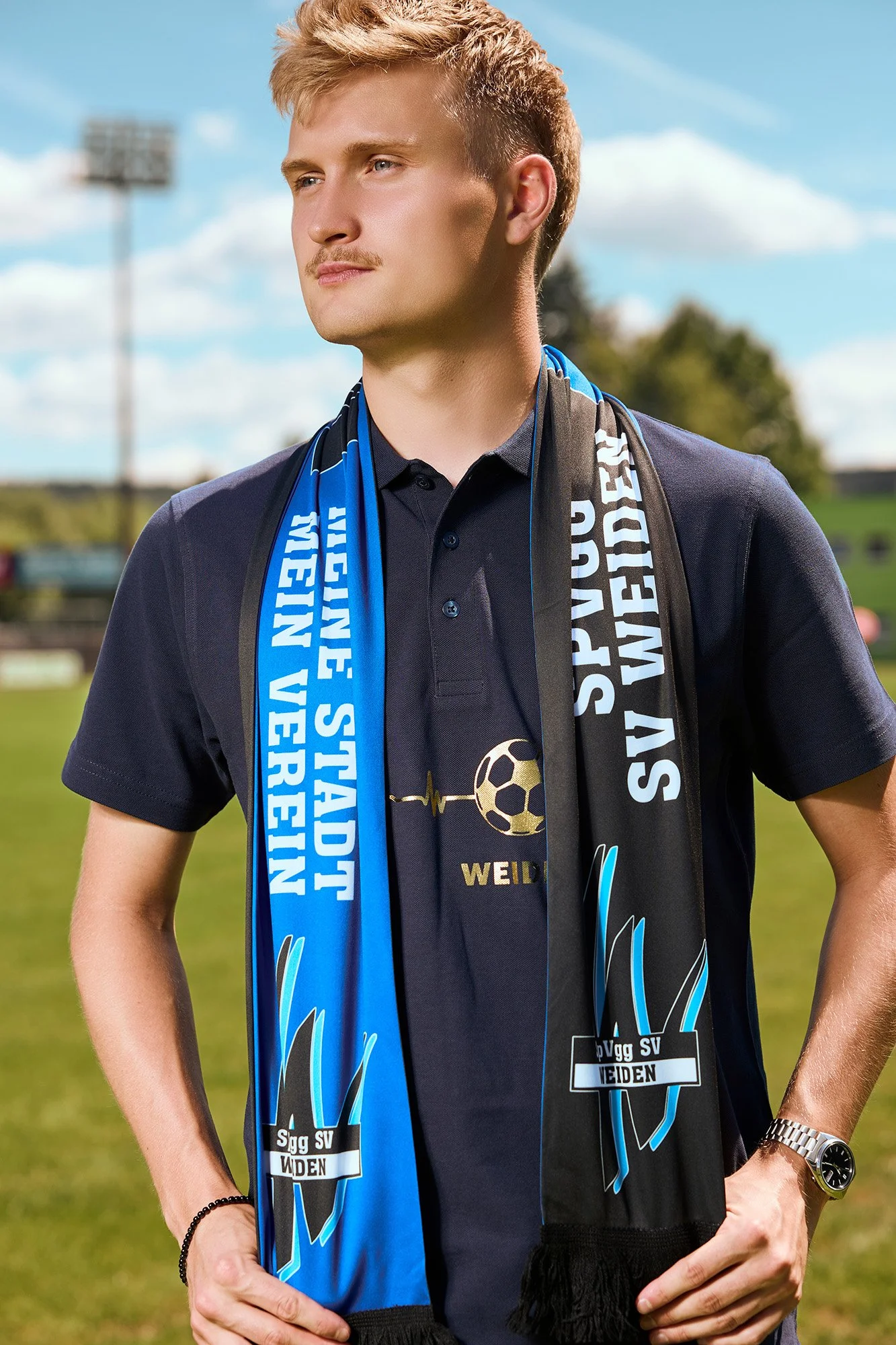 Mann mit blauen und schwarzen Fan-Schals und T-Shirt mit Fußballmotiven vor einem Fußballfeld, sonnigem Himmel und Bäumen im Hintergrund.