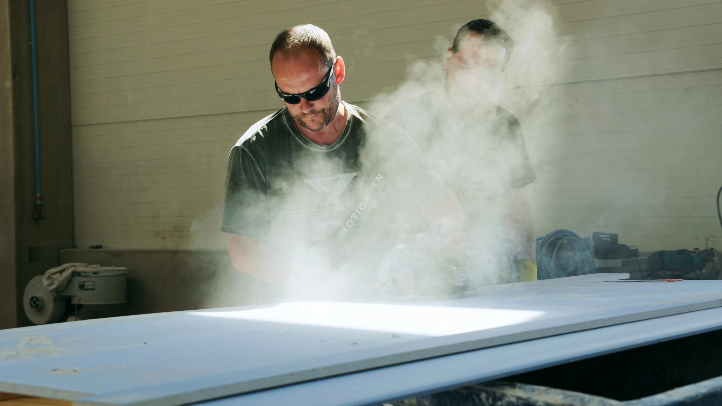 Zwei Männer arbeiten in einer Werkstatt, einer trägt Schutzbrille, während Rauch auf einer weißen Oberfläche aufsteigt.
