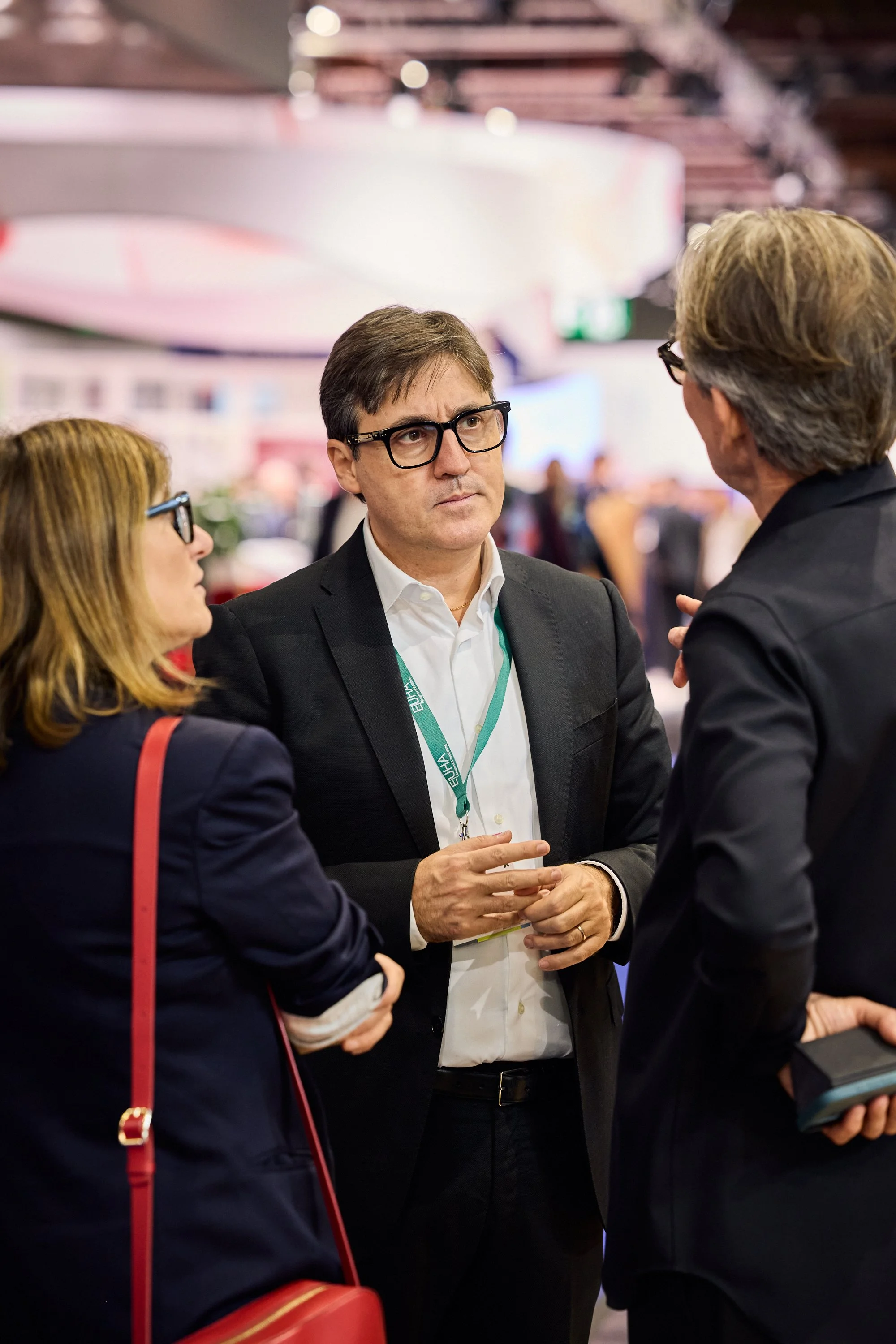 Drei Personen im Gespräch bei einer Konferenz oder Messe, im Hintergrund eine bunte Decke und Menschen.