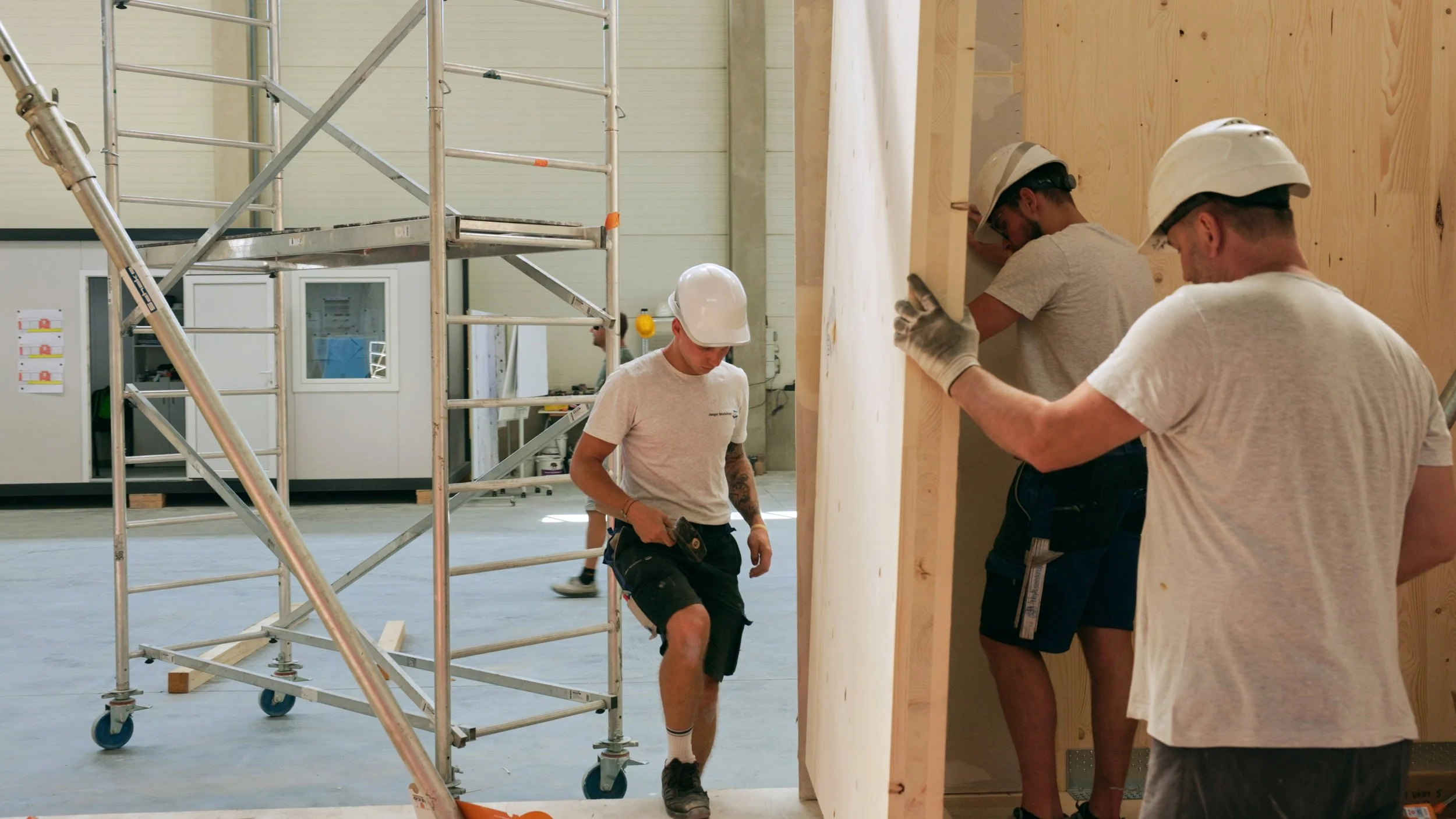 Drei Bauarbeiter tragen eine große Holzplatte in einer Bauhalle, alle mit Schutzhelmen und Arbeitskleidung, einer der Arbeiter hat einen Bohrer in der Hand.