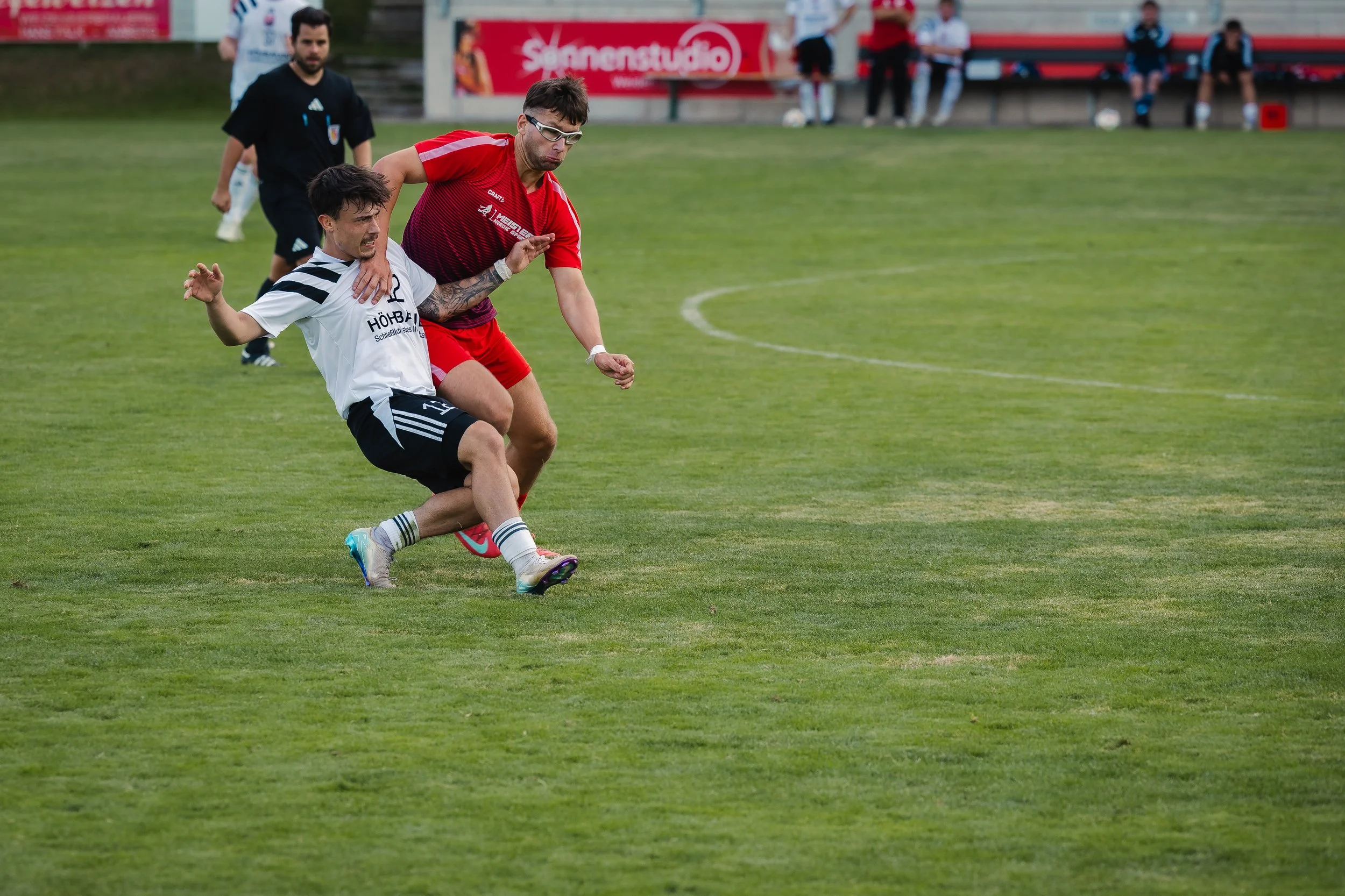 Zwei Männer kämpfen um den Ball während eines Fußballspiels auf dem Spielfeld, während Hintermannschaft und Zuschauer im Hintergrund zu sehen sind.