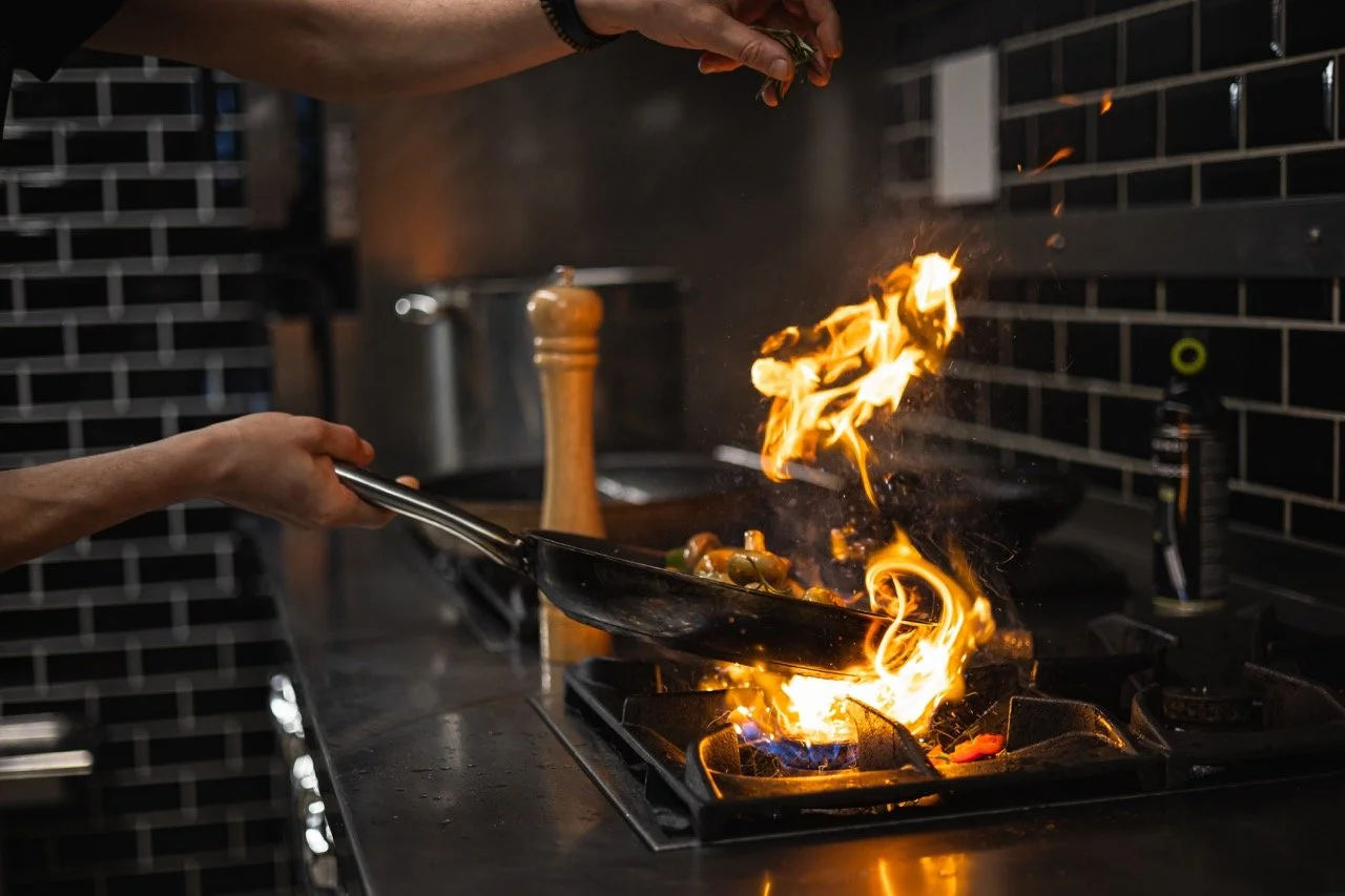 Koch in einer Küche, der in einer Pfanne flambiert, Flammen schlagen hoch, schwarze Fliesen im Hintergrund.