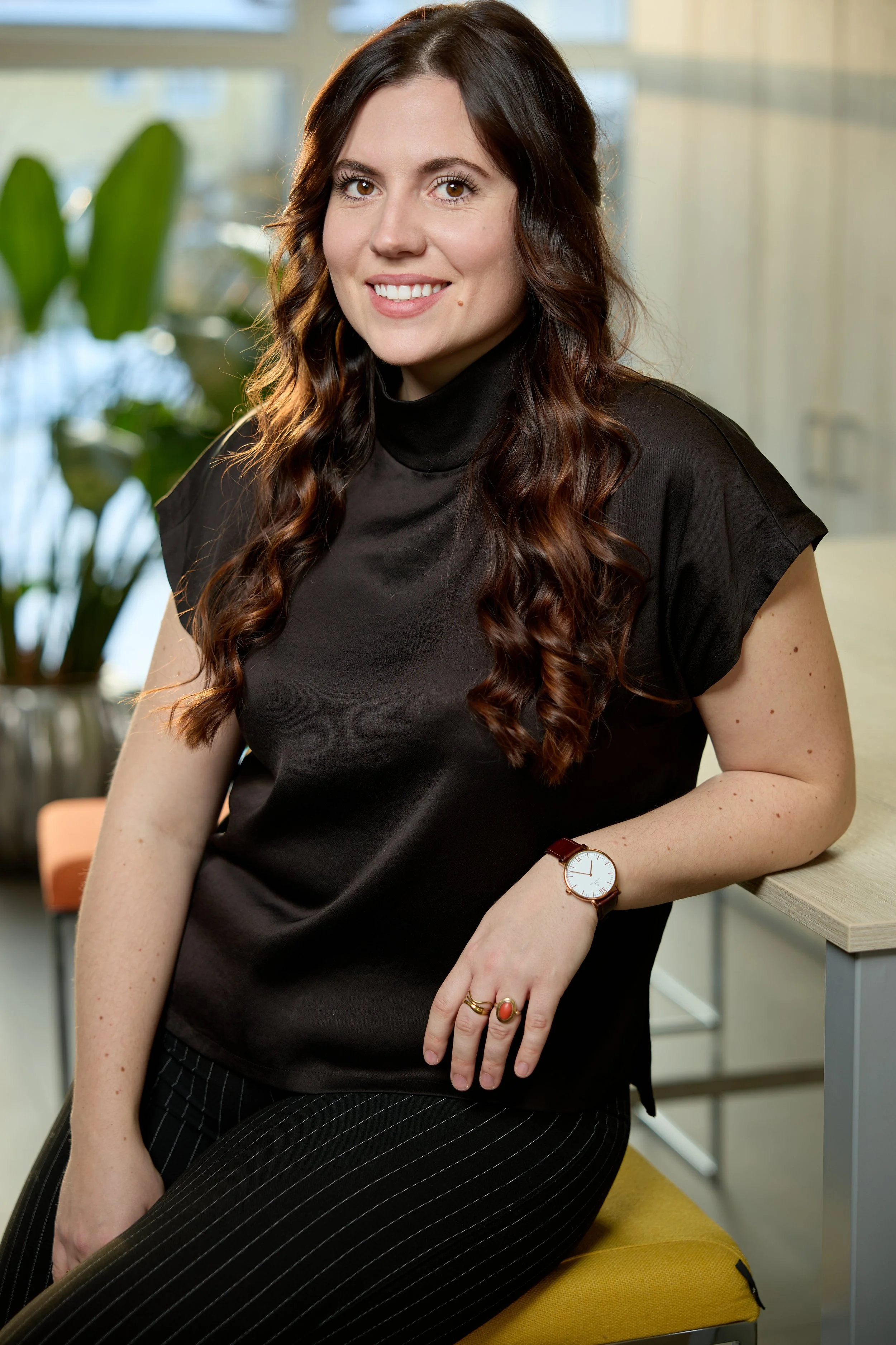 Business Portrait: Eine junge Frau sitzt in einem modernen Büro, trägt eine schwarze Bluse, hat lange, lockige braune Haare und trägt eine Armbanduhr und Ringe. Sie lächelt in die Kamera.