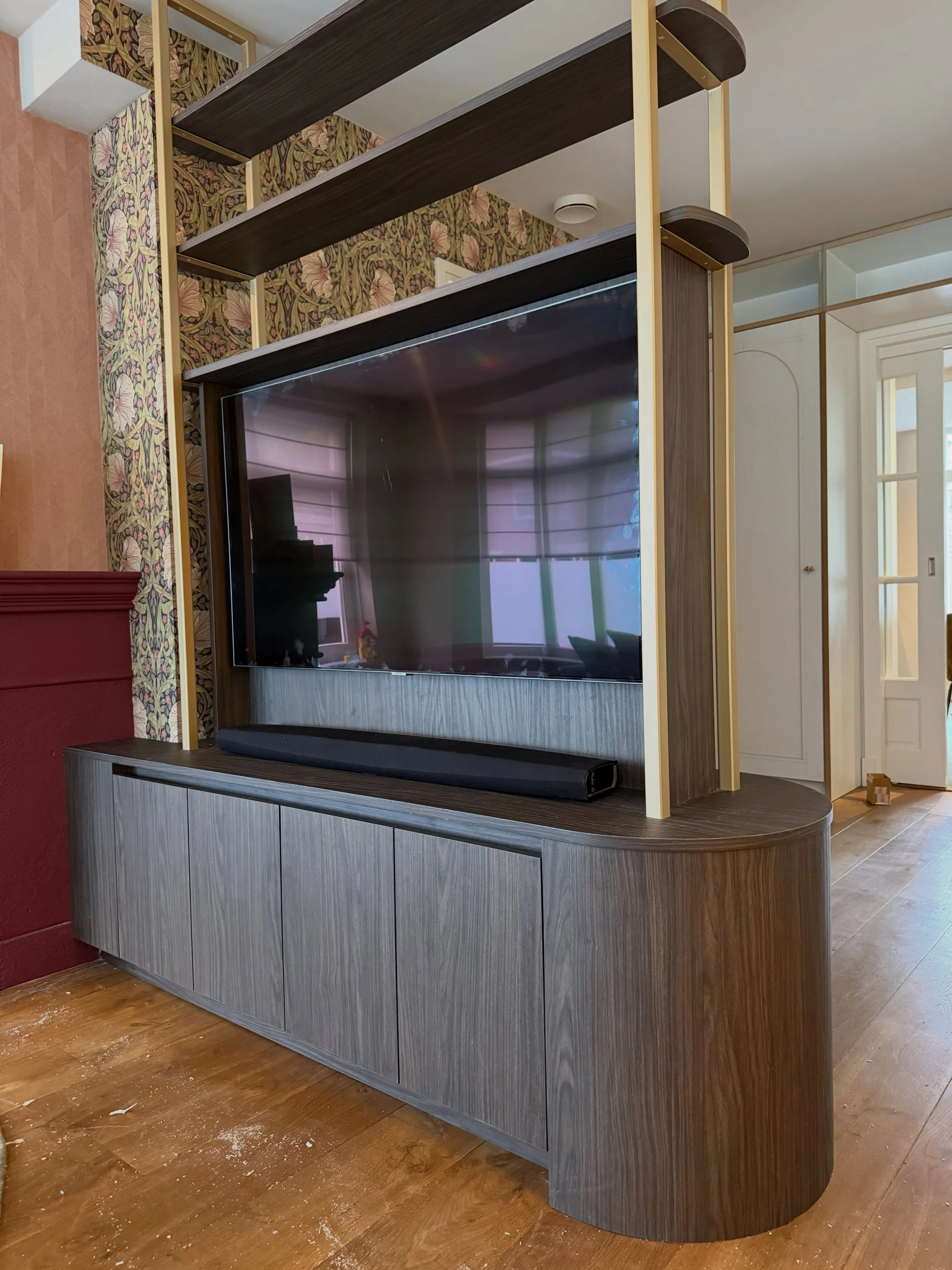 Maatwerk roomdivider met ge&iuml;ntegreerde tv. Afgewerkt met stalen staanders in een gespoten messingkleur in combinatie met een donkere houtlook, voor een stijlvolle en warme uitstraling.