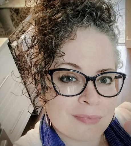 Close-up of a woman with curly hair, glasses, and earrings, smiling.