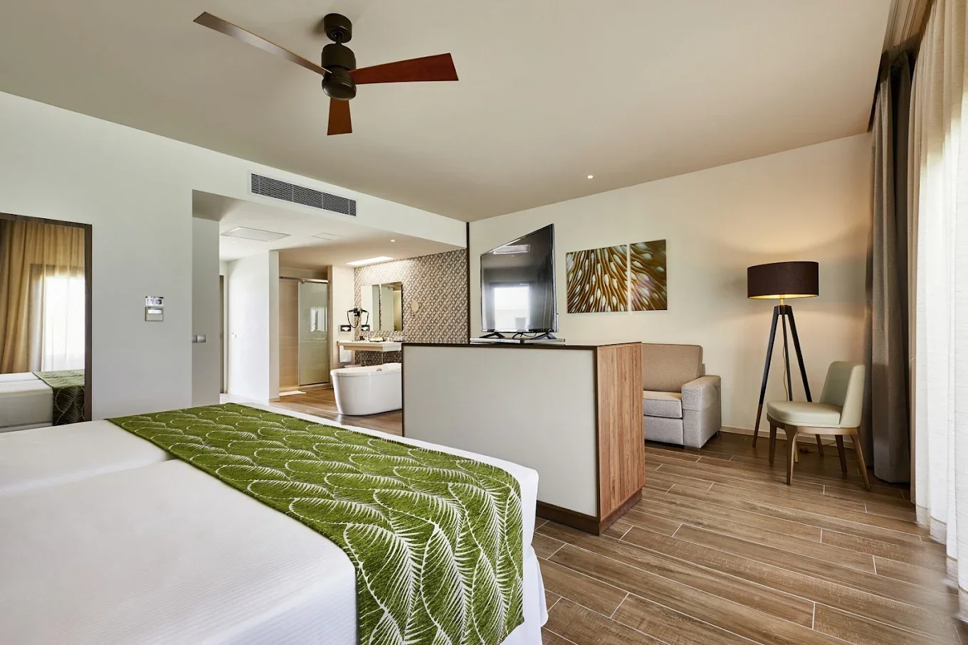 A hotel room with a bed, green leaf-patterned blanket, wood flooring, television, sitting area with armchair and floor lamp, and a bathroom with a bathtub visible in the background.