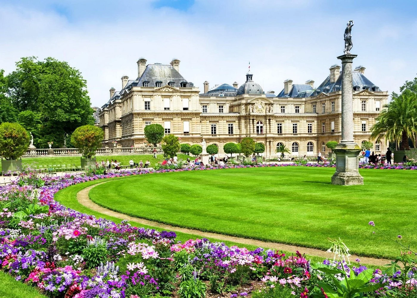 A historic mansion with a garden in front, featuring green grass, colorful flowers, and neatly trimmed bushes; a fountain with a tall column and statue is visible in the foreground.