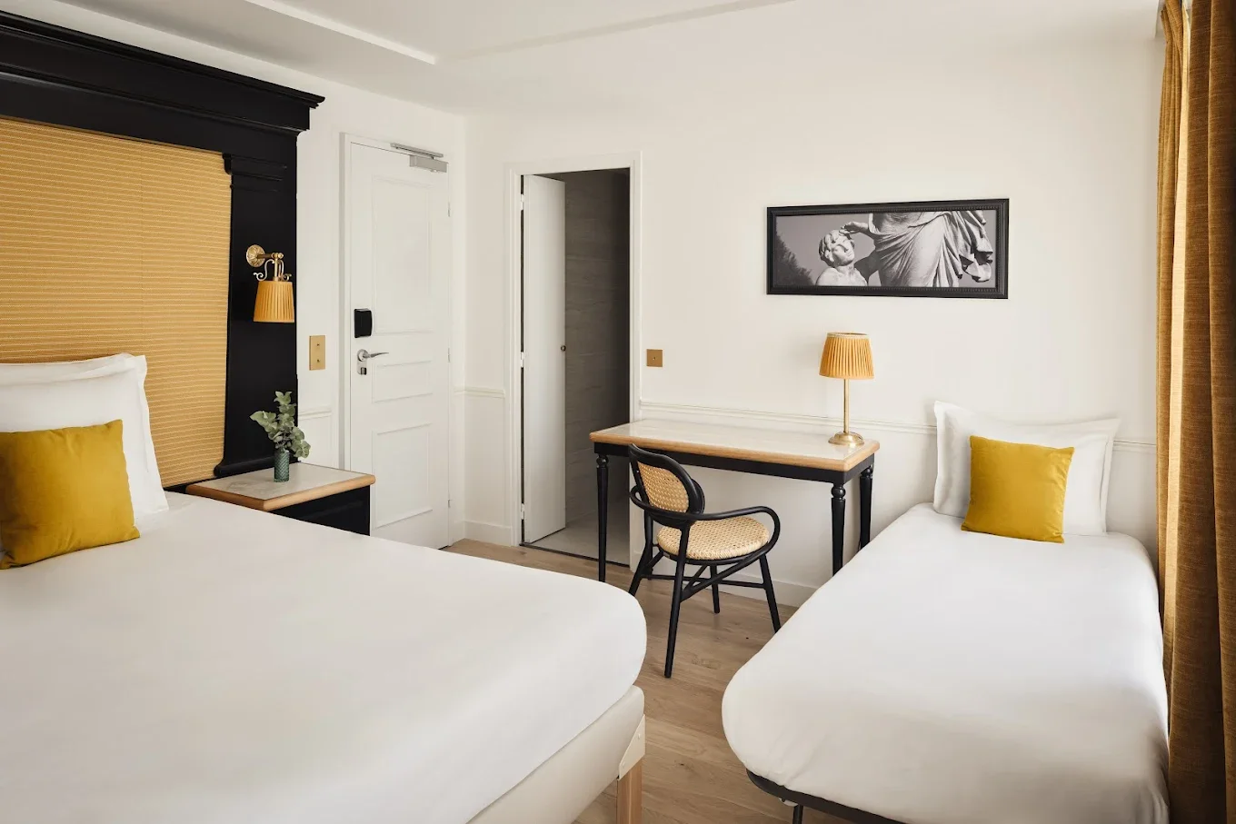 Hotel room with two white beds, yellow pillows, a black and wooden headboard, a small side table with a plant, a desk with a lamp, a chair, a black and white wall art, and curtains.